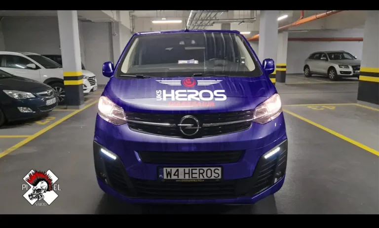Niebieski van Opla na krytym parkingu, zwrócony przodem do kierunku jazdy. Na masce widnieje napis "UKS HEROS WARSZAWA" z pogrubionymi biało-czerwonymi literami i czerwonym logo w kształcie byka. Na tablicy rejestracyjnej widnieje napis "W4 HEROS". W lewym dolnym rogu znajduje się logo z czaszką i napisem "PIXEL AUTO".